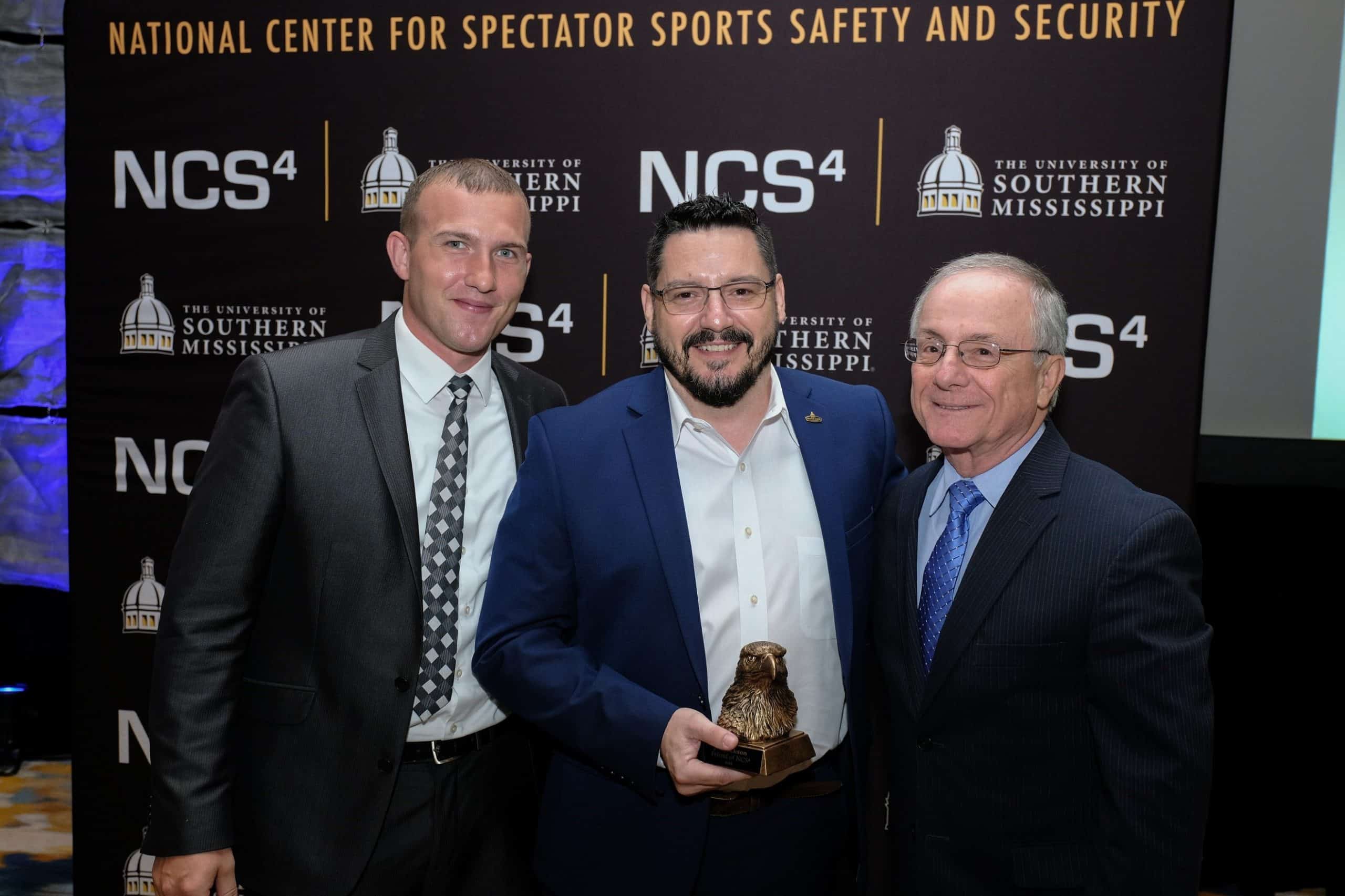 Three men in suits smile; the center holds an eagle award. NCS4 and USM logos are in the backdrop.