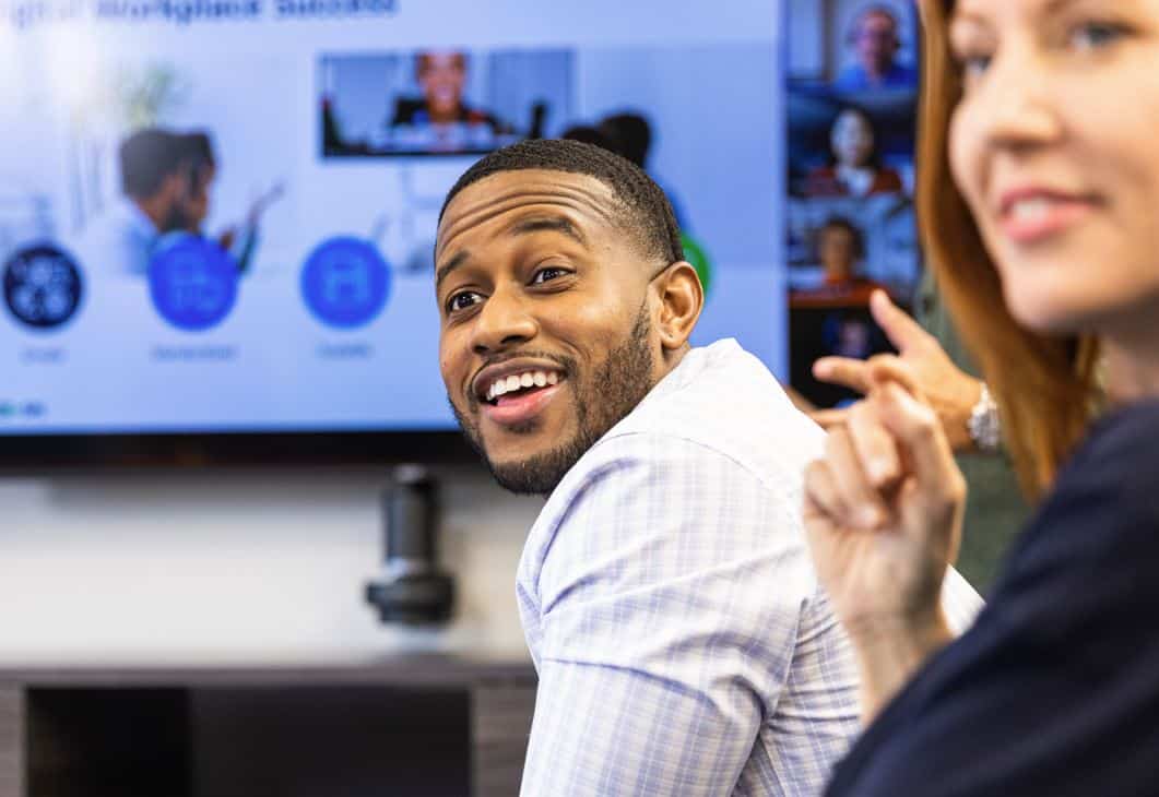 Man in checkered shirt smiles back at colleague in meeting room, large screen with video call and graphics in background.