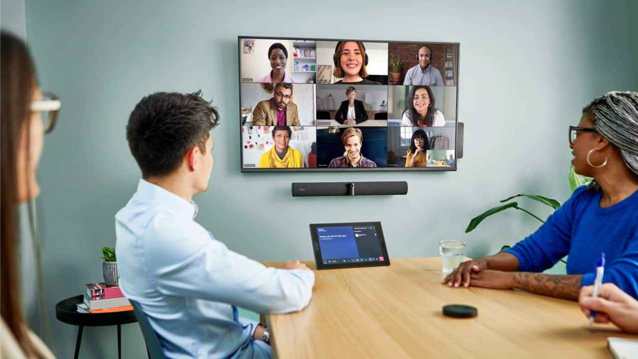 Four people at a conference table on a video call with eight others, tablet and speaker on the table, plant in the background.