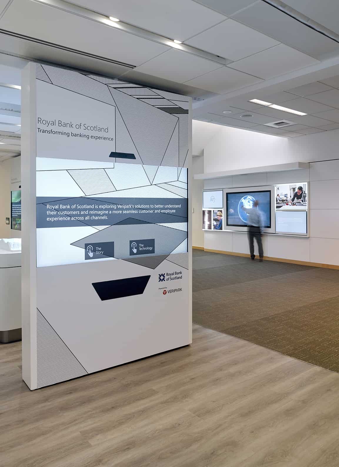 Modern bank with digital displays, wood flooring, exhibits; sign reads Royal Bank of Scotland: Transforming banking experience.