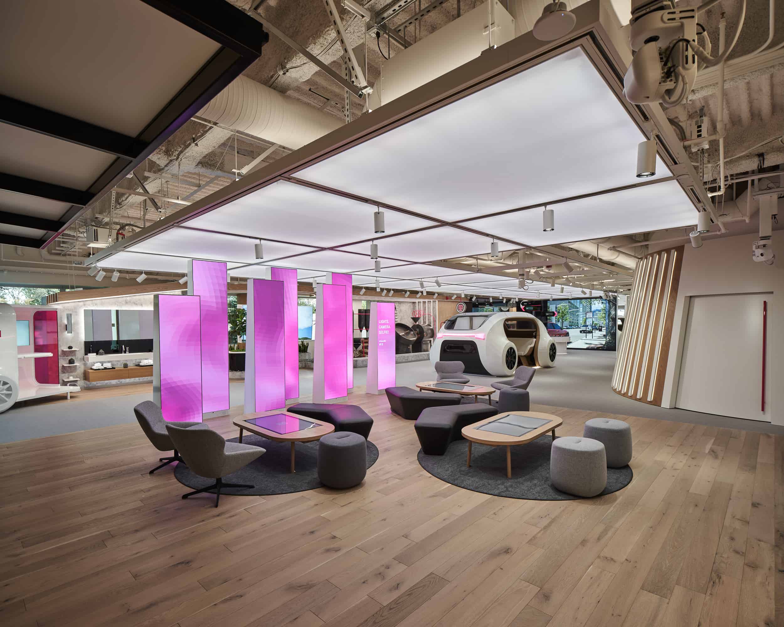 Modern showroom with futuristic car, vibrant pink lights, dark chairs, round tables, and warm wooden flooring.