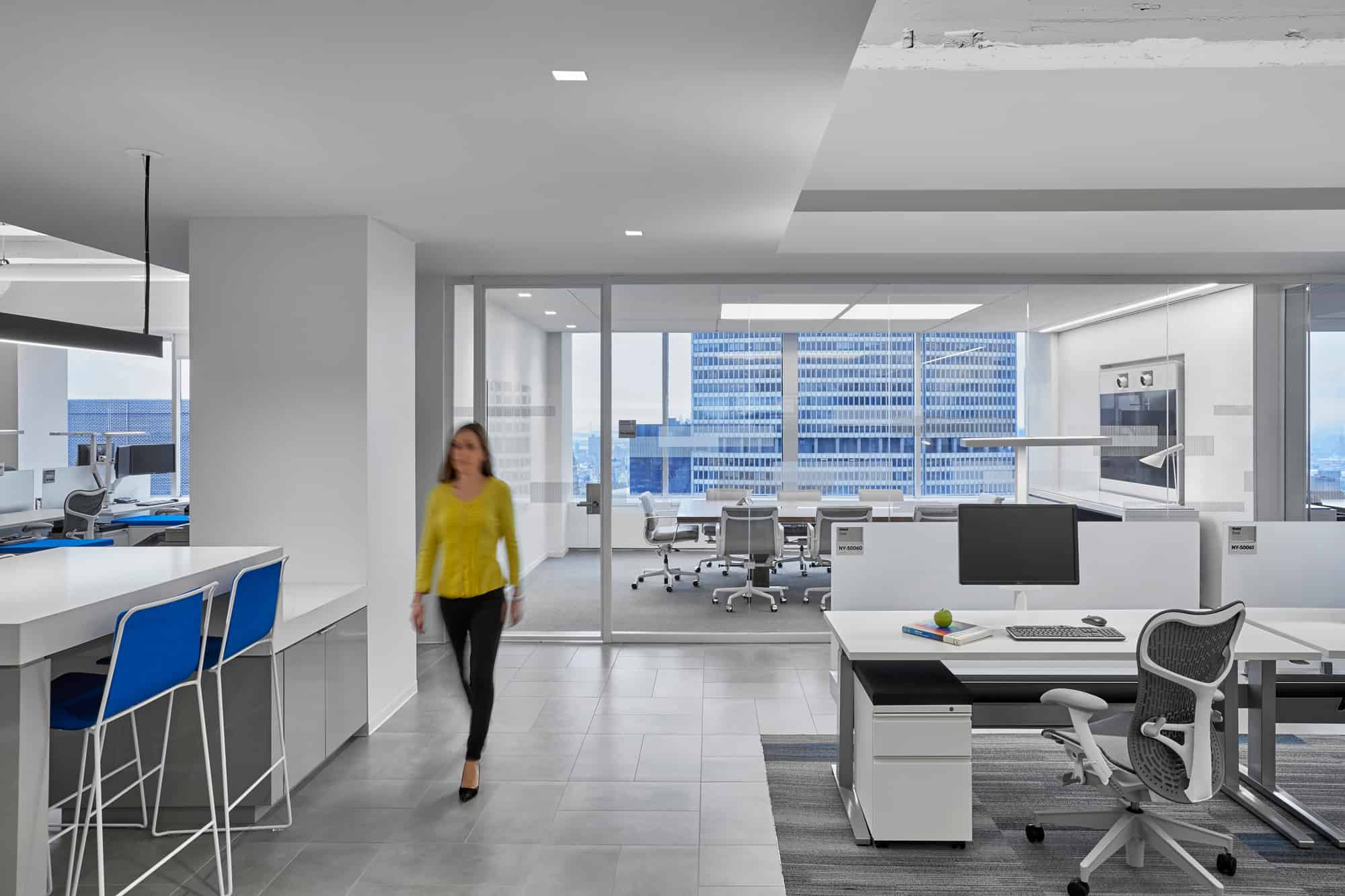 Modern office: bright, minimalist; person in yellow walks by desks. Blue seating area in front, cityscape visible through large window.