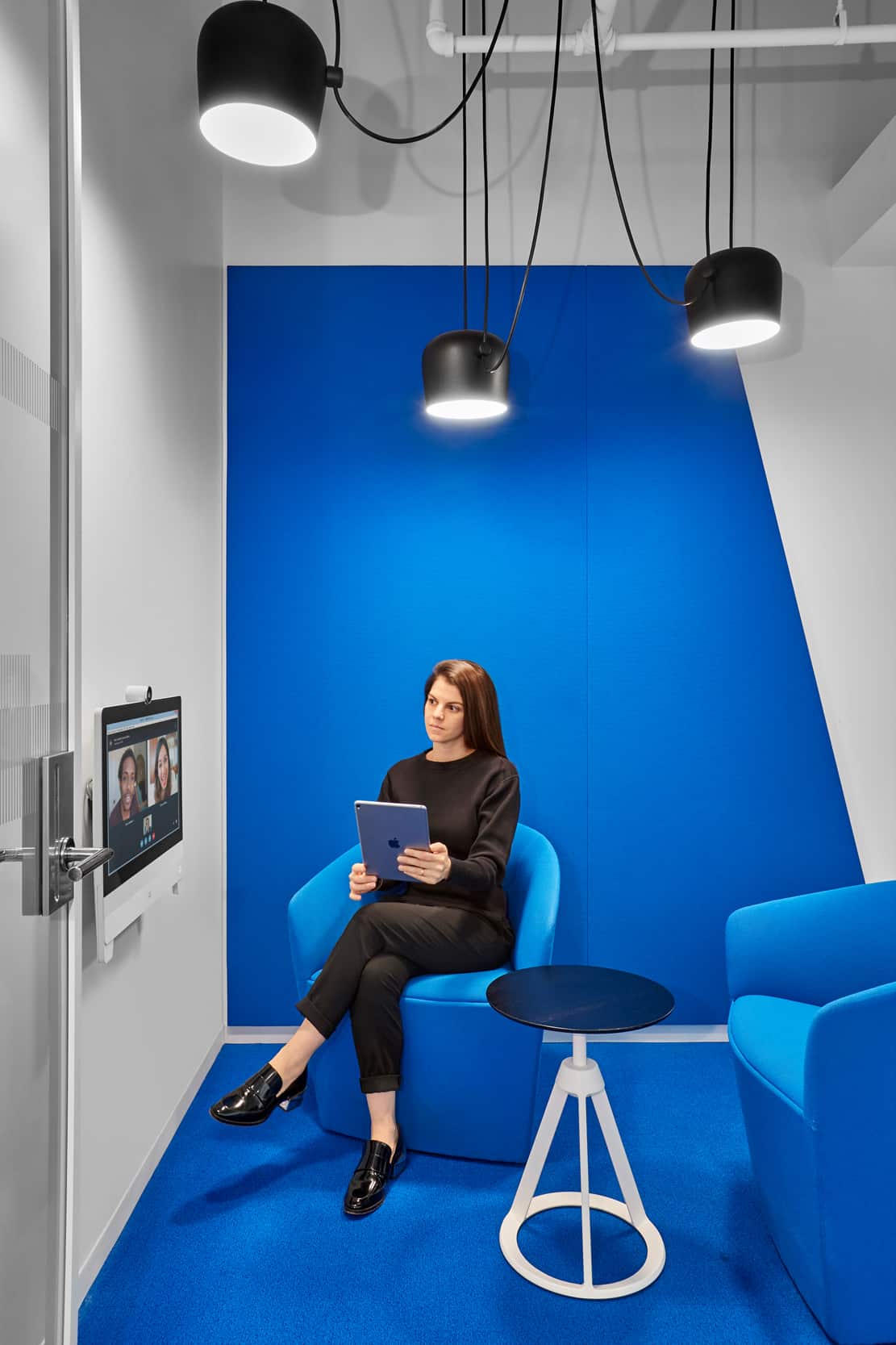 Woman in modern office with blue walls, holding tablet; monitor shows video call. Ceiling lights above, small round table nearby.