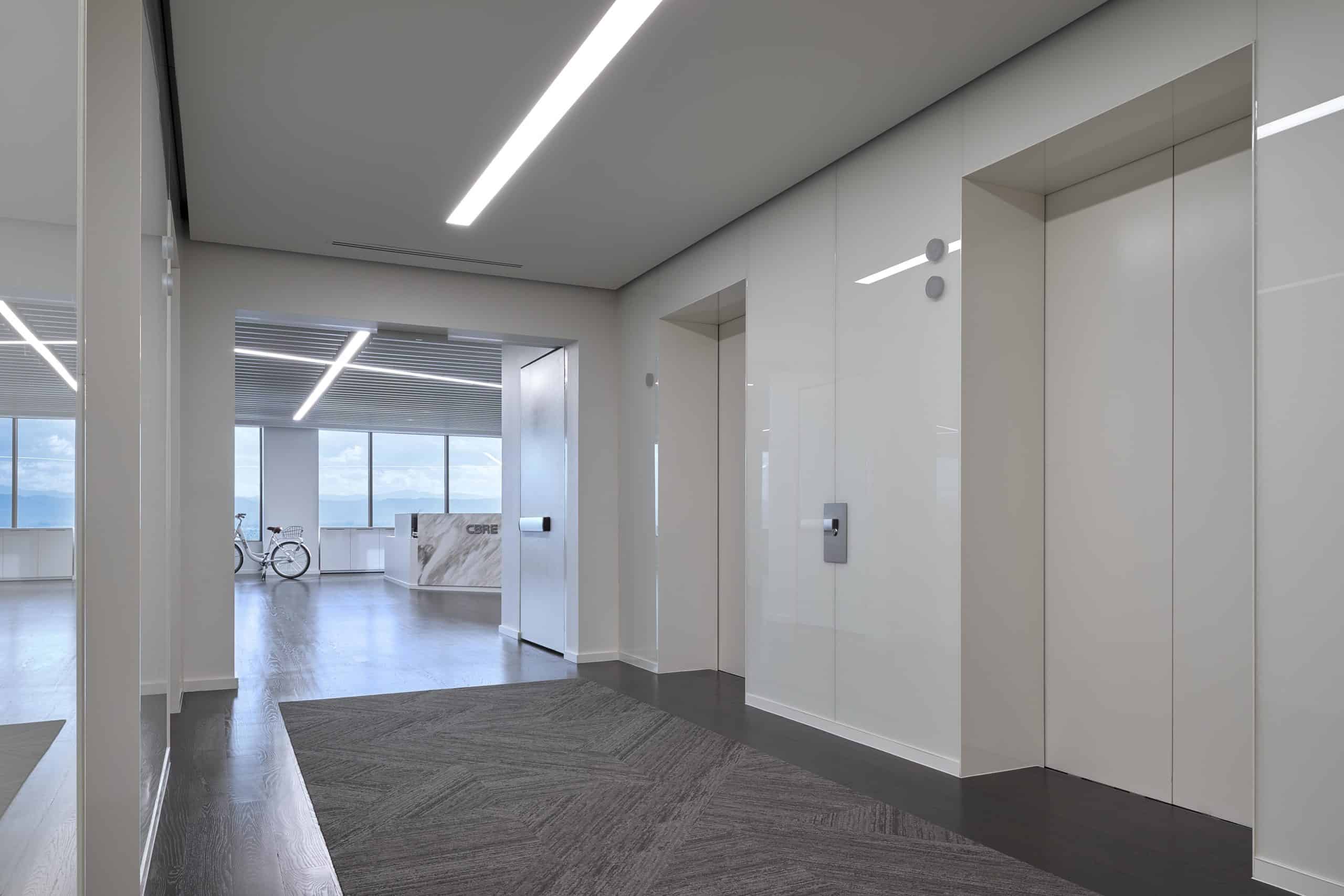 Modern hallway: white walls, wood floors, two elevator doors right, large windows ahead with sky view; bike parked by windows.