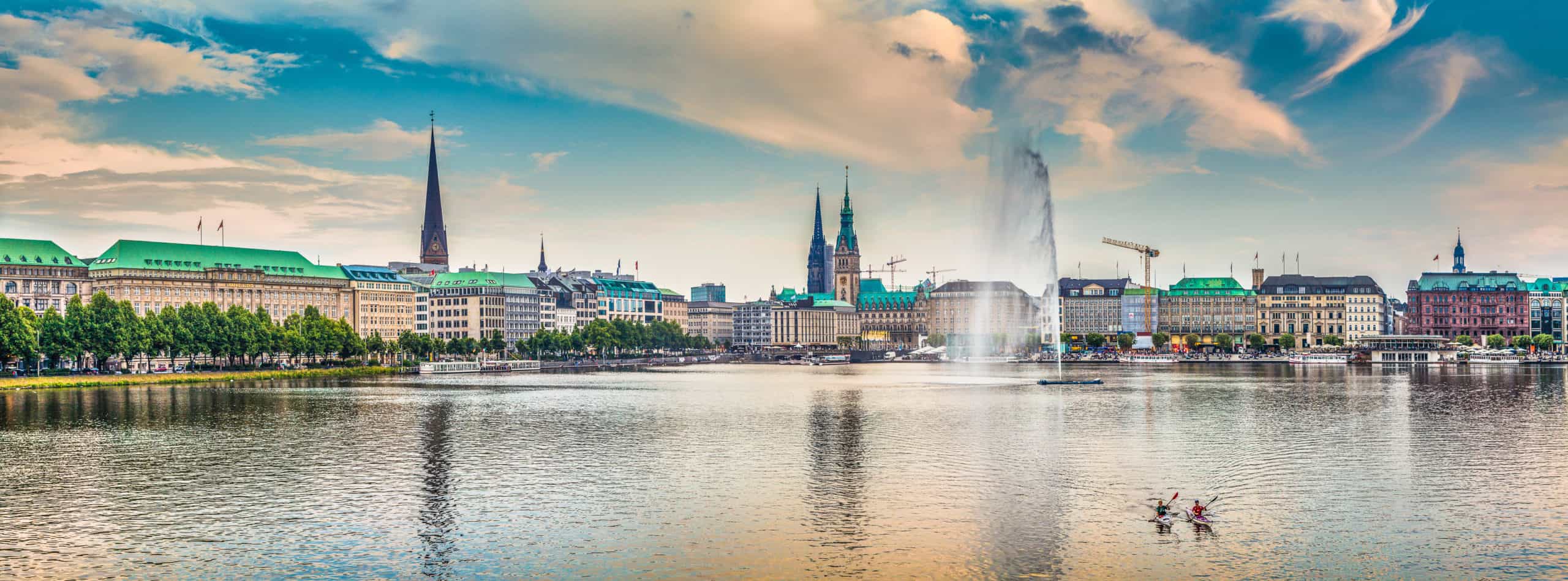 Hamburg skyline: Binnenalster lake, church spires, modern buildings. Fountain sprays; kayakers paddle under dramatic clouds.