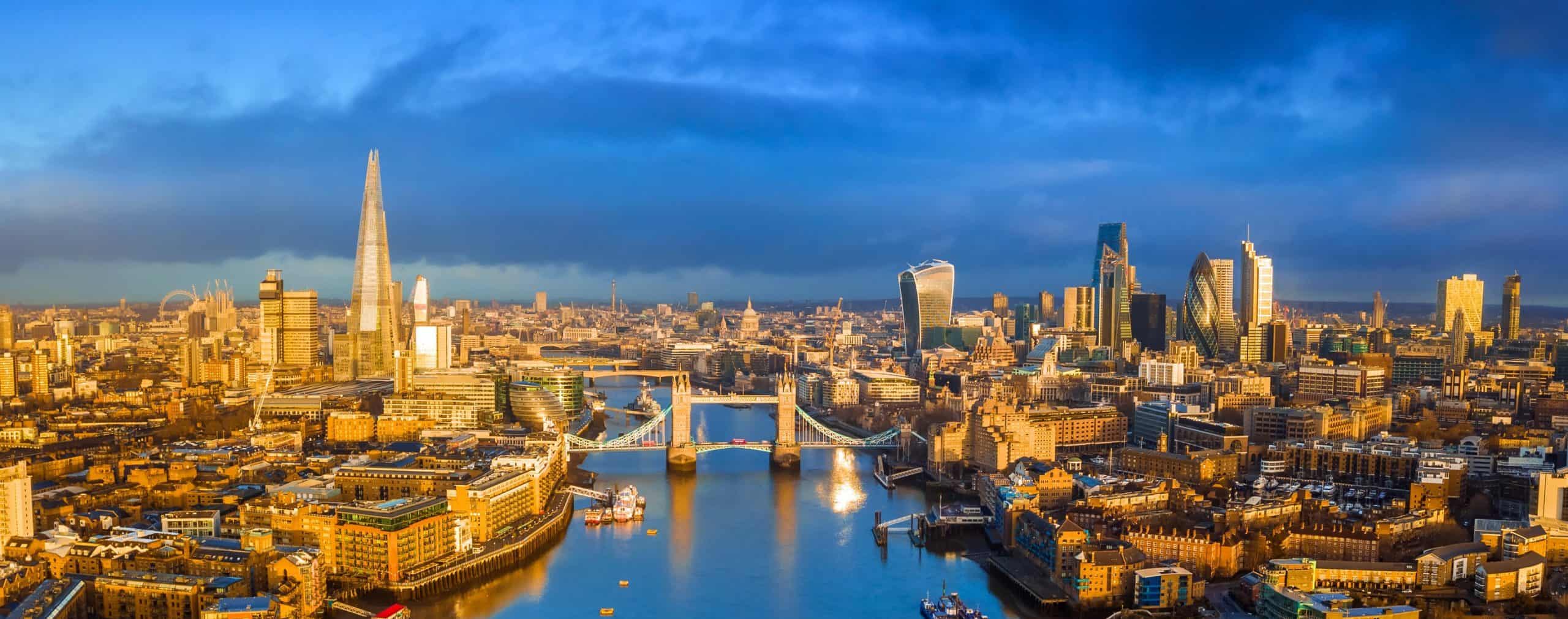 Aerial view of River Thames, Tower Bridge, the Shard, and Walkie Talkie under a sunny sky with clouds. Skyscrapers bask in warm light.