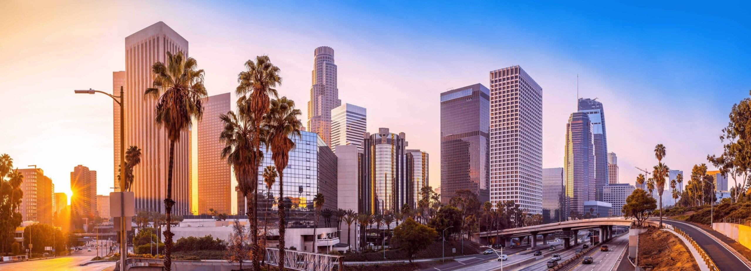 City skyline with skyscrapers & palm trees at sunset; warm glow over buildings, freeway bustling with cars below.