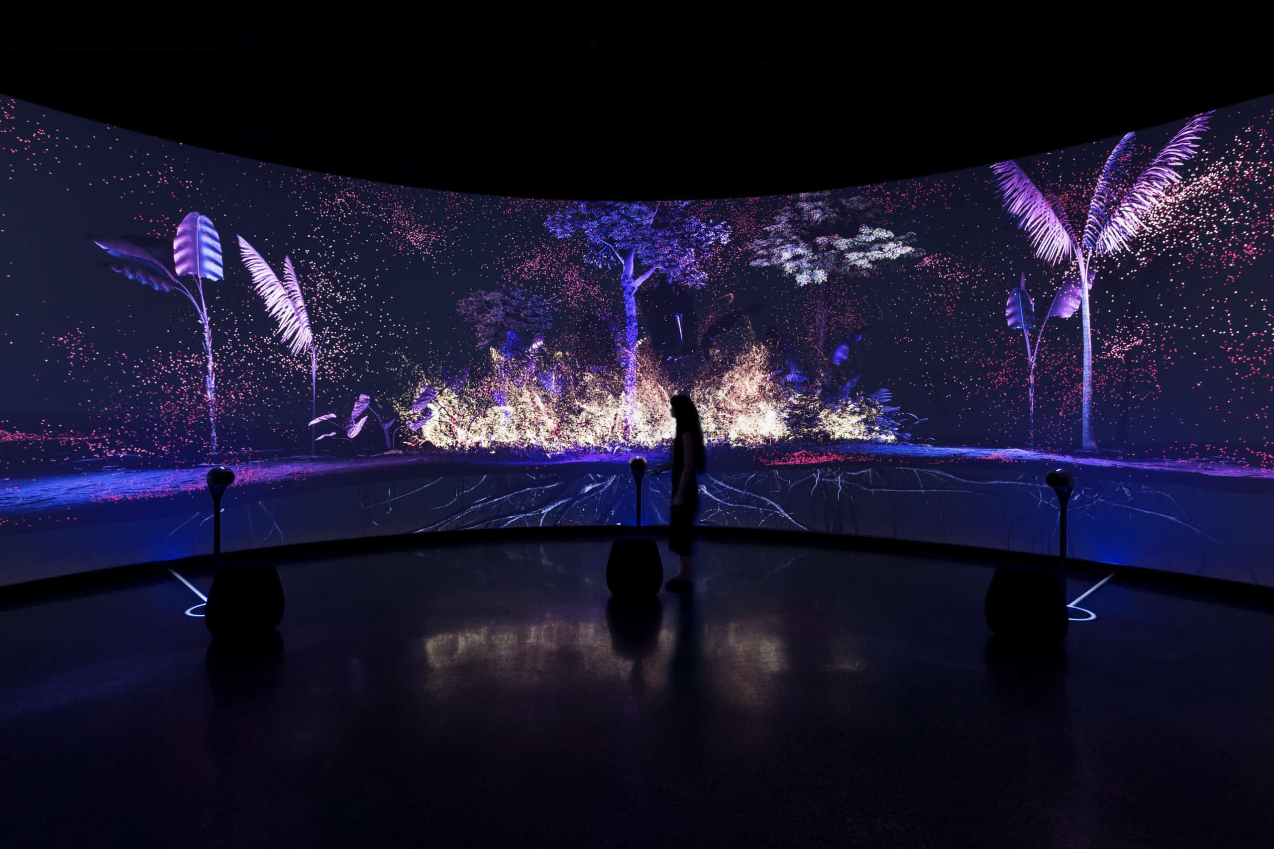 A person stands before a curved screen showing vibrant digital art with abstract trees, in a dim room with surrounding speakers.