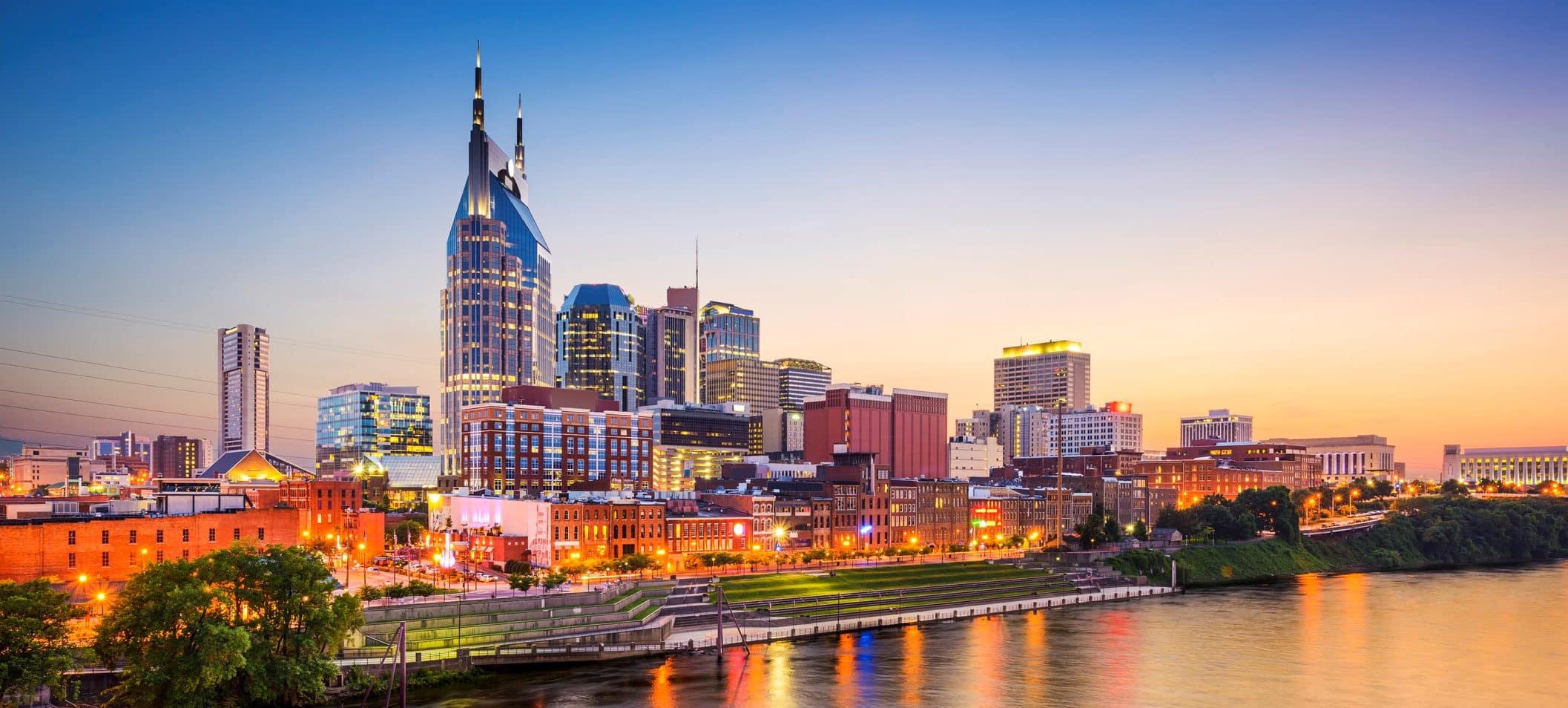 Nashville skyline at dusk with the AT&T Building reflected in a river, as city lights glimmer under a blue-to-orange sunset sky.