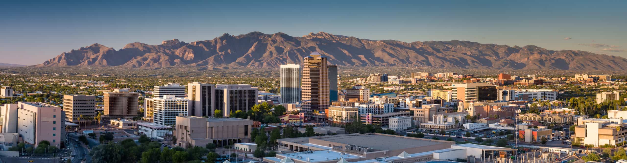 Cityscape with tall offices, residential areas, rugged mountains, and clear blue sky in a panoramic view.