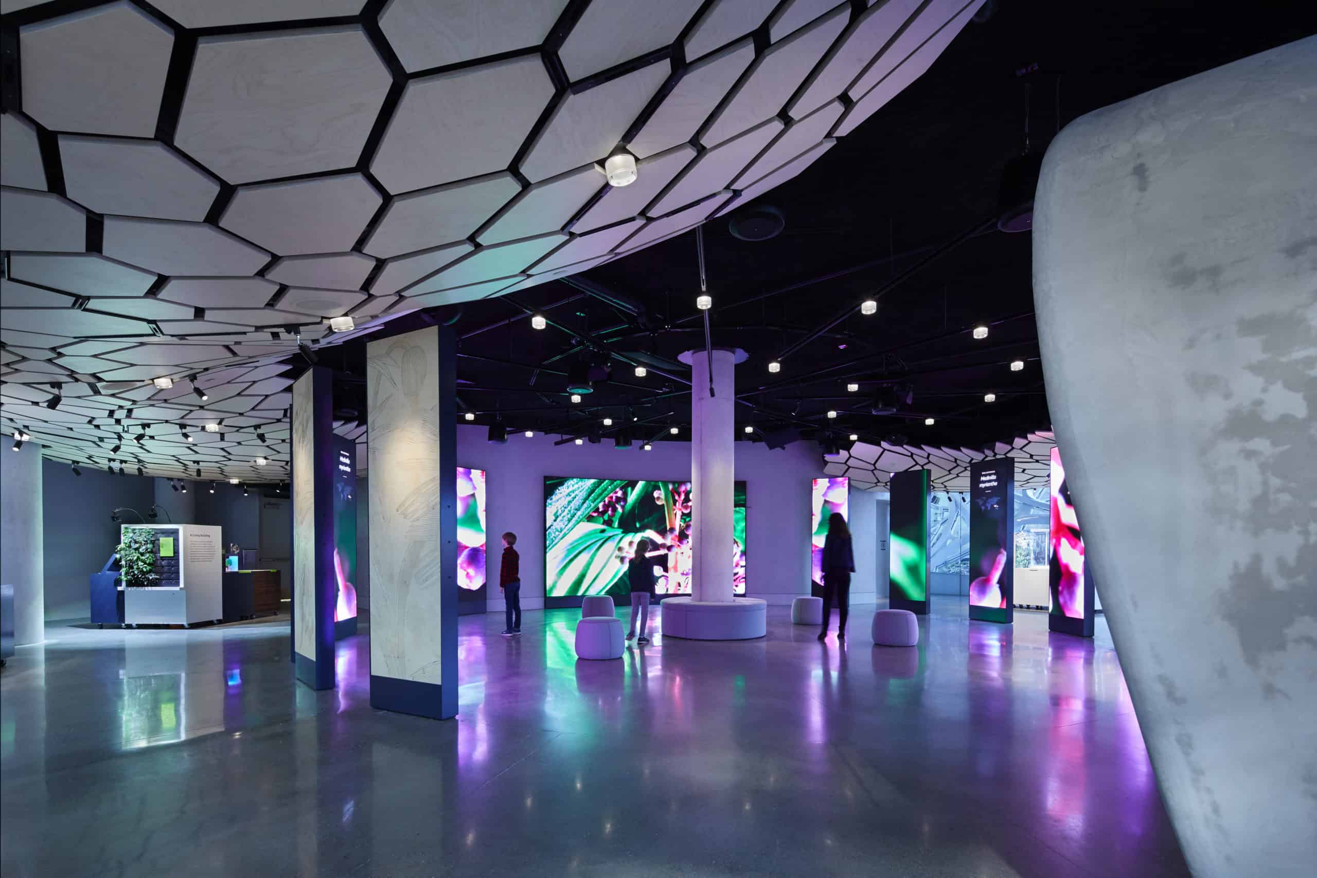 Modern space with hexagonal ceiling panels, vibrant digital displays, glossy floor, columns, and stools for a sleek, futuristic vibe.