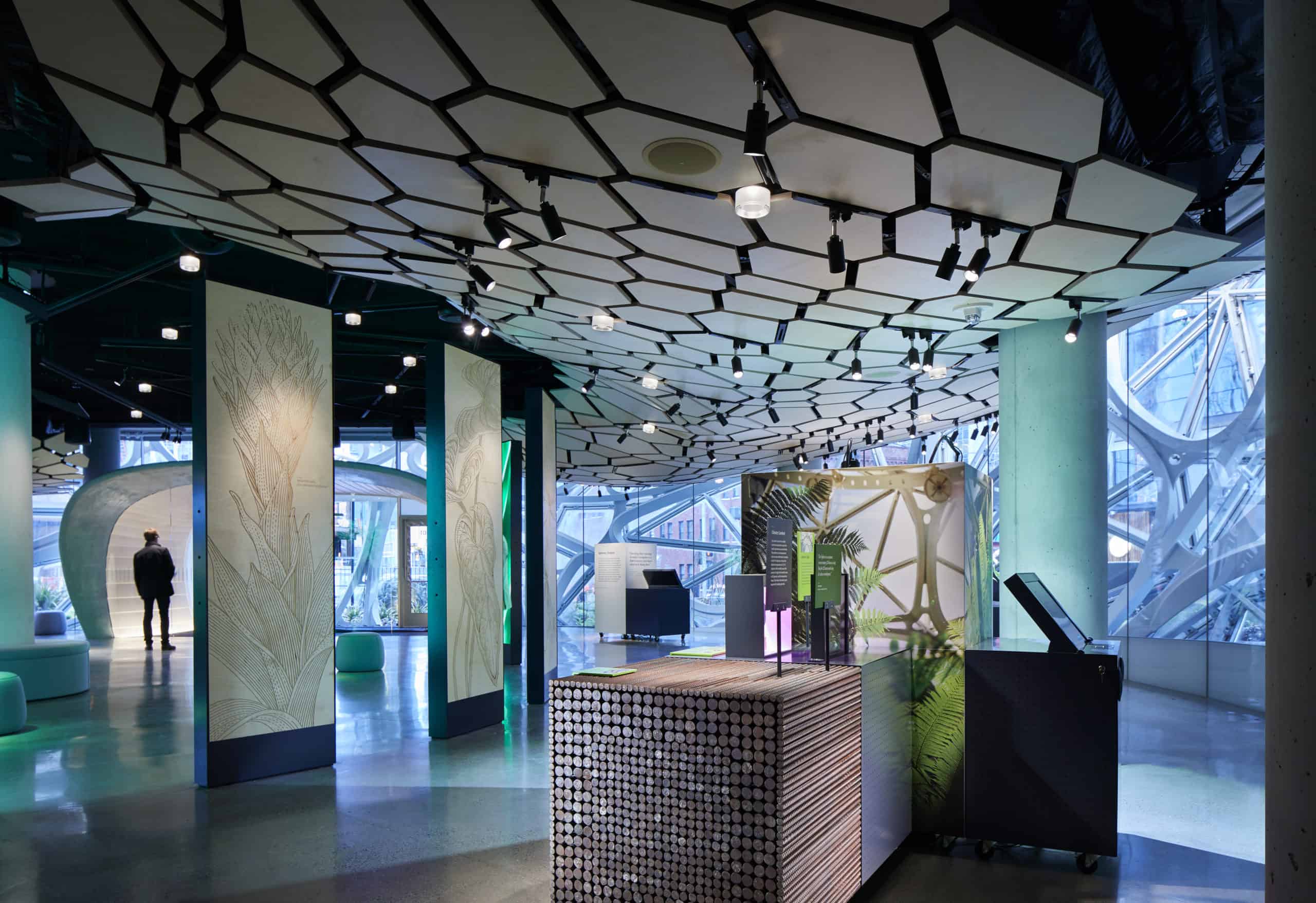 Modern interior with geometric ceiling, spotlights, abstract wall panels, glass windows, unique desk with plants; person in distance.