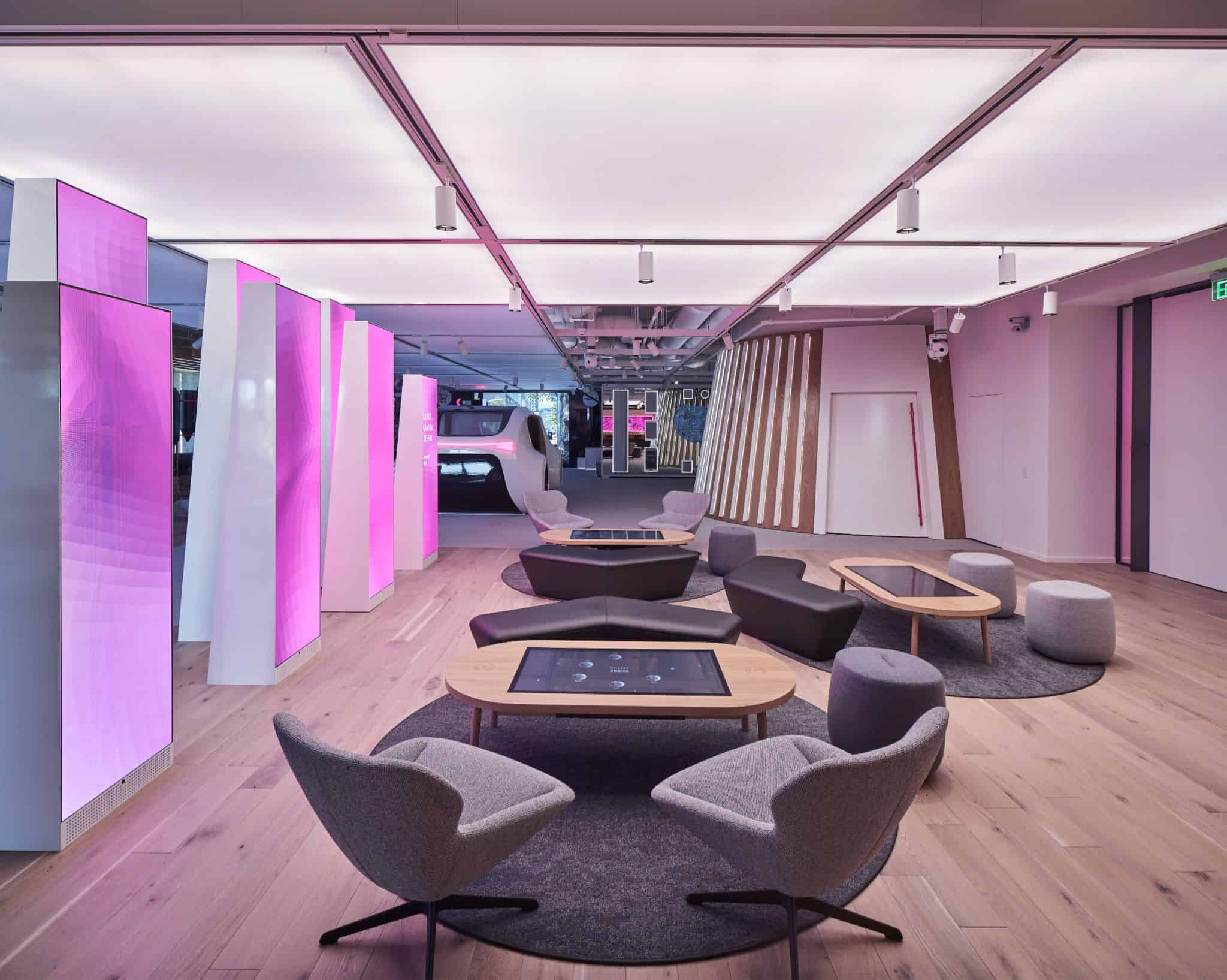 Sleek office lobby: Bright lights, futuristic pink/white columns, gray chairs, round tables on rugs, light wood floors.