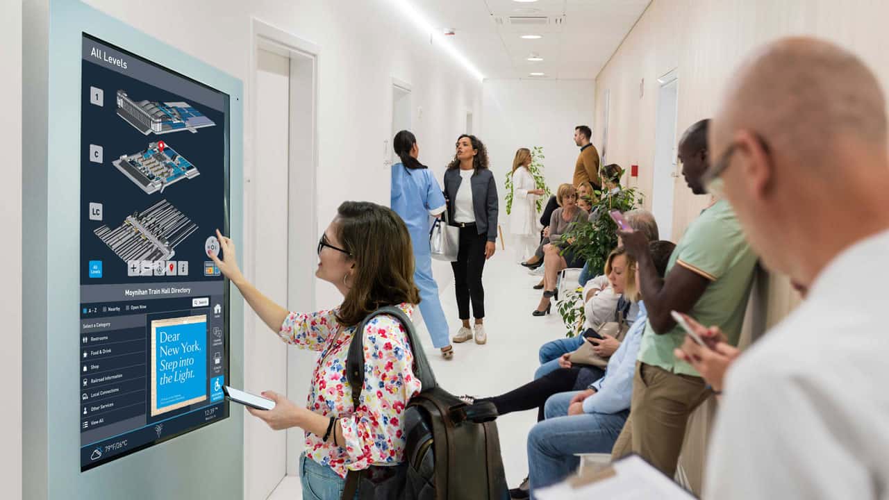A woman in glasses uses a touchscreen map on the wall; others sit nearby with devices, in a modern, brightly lit hallway.
