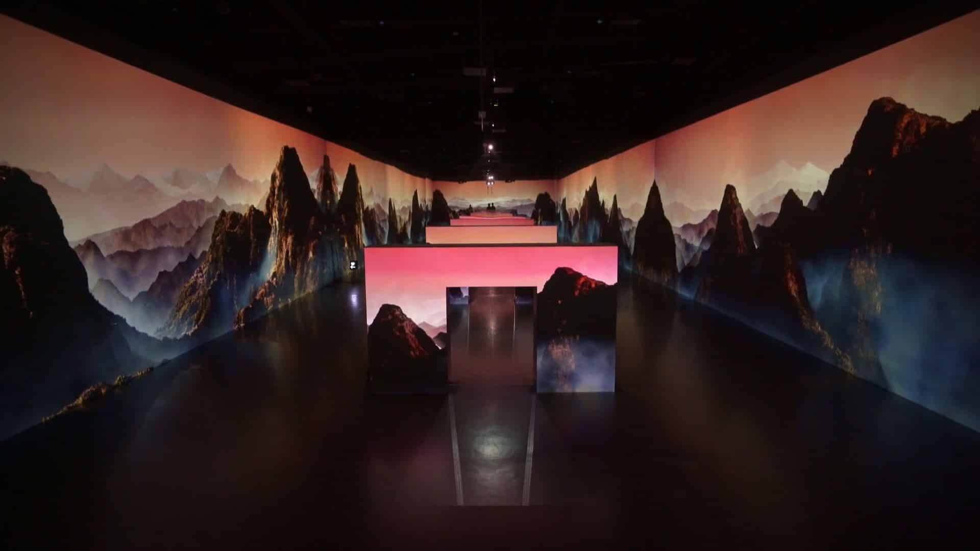 A dimly lit room with large screens shows a panoramic mountain landscape at sunset, creating an immersive pathway.