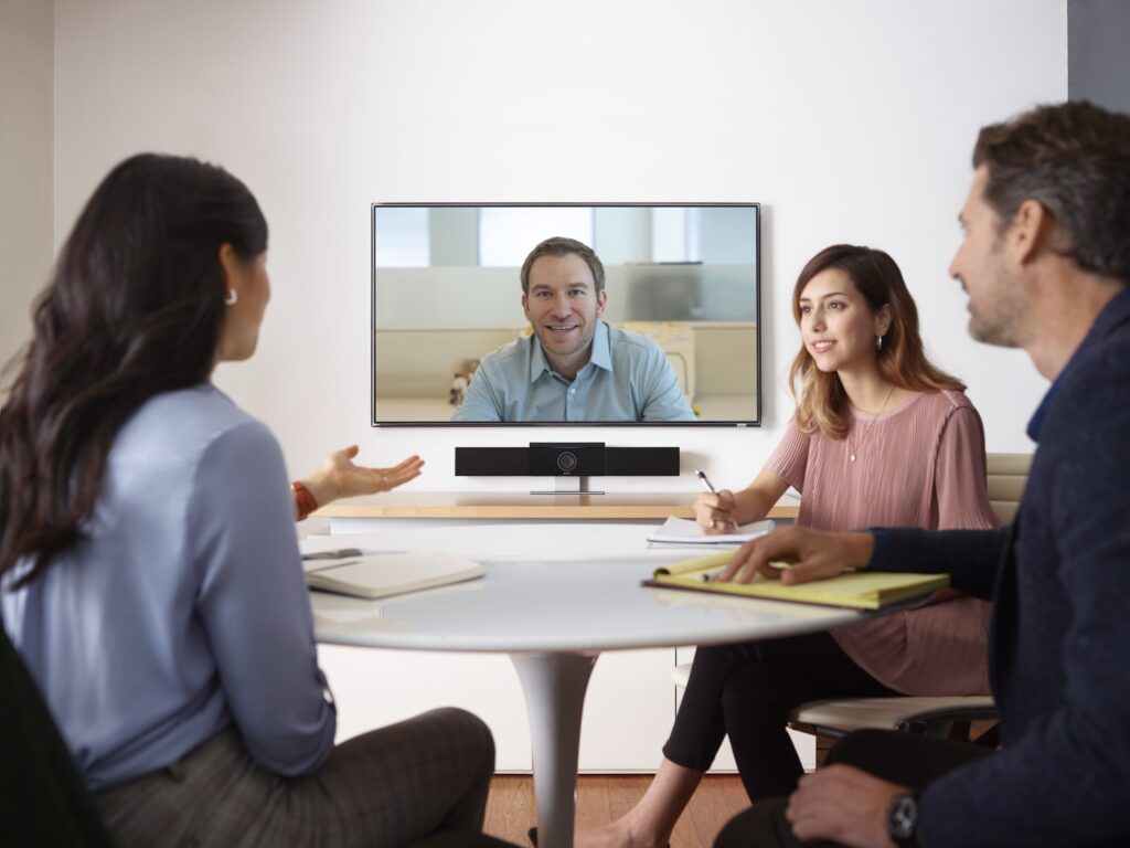 Four people in a video conference: three at a round table with notebooks/laptop, one on screen.